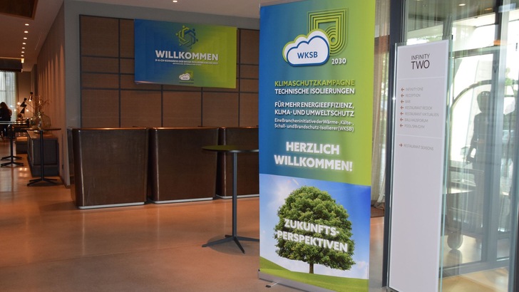 Ein Banner in einer Lobby, das für technische Dämmung zur Verbesserung der Energieeffizienz von Gebäuden wirbt. Im Hintergrund sind Schilder und Tische zu sehen, die auf einer Veranstaltung zum Thema innovative Dämmstoffe und Normen in der Gebäudetechnik hinweisen. - © RM Rudolf Müller Medien GmbH & Co. KG Ein Banner in einer Lobby, das für technische Dämmung zur Verbesserung der Energieeffizienz von Gebäuden wirbt. Im Hintergrund sind Schilder und Tische zu sehen, die auf einer Veranstaltung zum Thema innovative Dämmstoffe und Normen in der Gebäudetechnik hinweisen. - © RM Rudolf Müller Medien GmbH & Co. KG