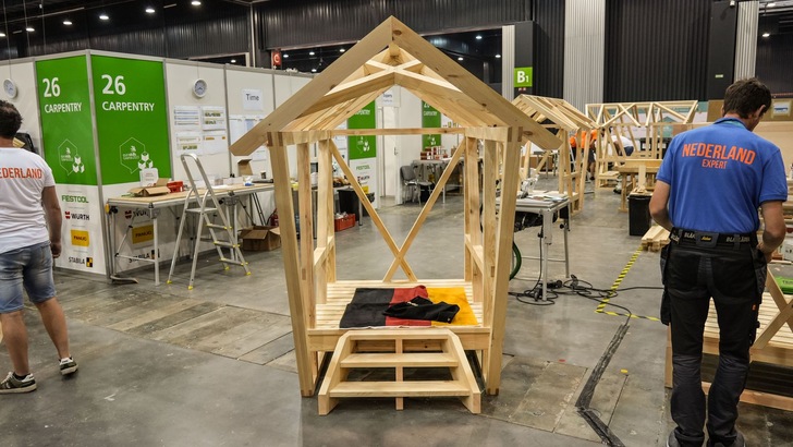 „Demonstration eines innovativen Dämmstoffs im Rahmen eines Holzhauses in einer Werkstatt, mit Fachleuten im Hintergrund, die an Dämmtechniken für Wärme-, Kälte- und Schallschutz arbeiten. - © Zentralverband des Deutschen Baugewerbes (ZDB)