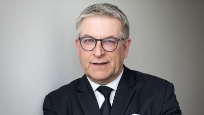 Mann mit grauem Haar und Brille, Anzug und Krawatte, posiert vor grauem Hintergrund. - © Achim Aulke
