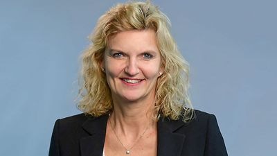 Lächelnde Person mit lockigem blondem Haar, die einen schwarzen Blazer vor einem schlichten blauen Hintergrund trägt. - © Claudia Westermann