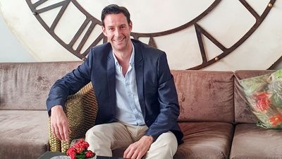 Mann im blauen Blazer sitzt auf einem Sofa vor einem großen Zifferblatt. - © Markus Biland
