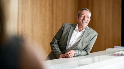 Lächelnder Mann im grauen Anzug lehnt an einer weißen Theke in einem holzgetäfelten Raum. - © Thomas Graber