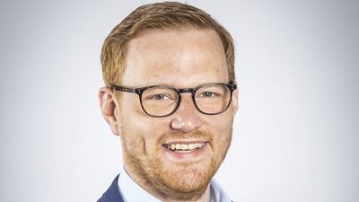 Ein Mann mit Brille und kurzen roten Haaren im Anzug lächelt vor einem neutralen Hintergrund. - © Tobias Erpenbeck
