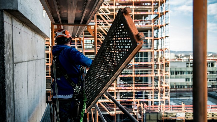 Ein Bauarbeiter in Sicherheitskleidung trägt auf einer Baustelle ein großes Metallgitter. Im Hintergrund ist ein teilweise errichtetes Gerüst sichtbar.