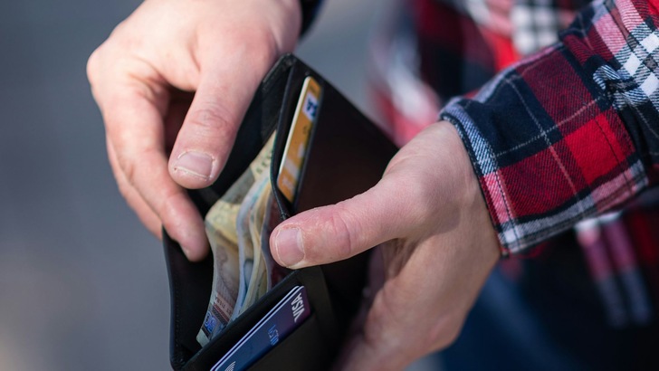 Bild einer Person, die eine Geldbörse hält und mit den Fingern Banknoten entnimmt. Die Person trägt ein kariertes Hemd und steht im Freien.