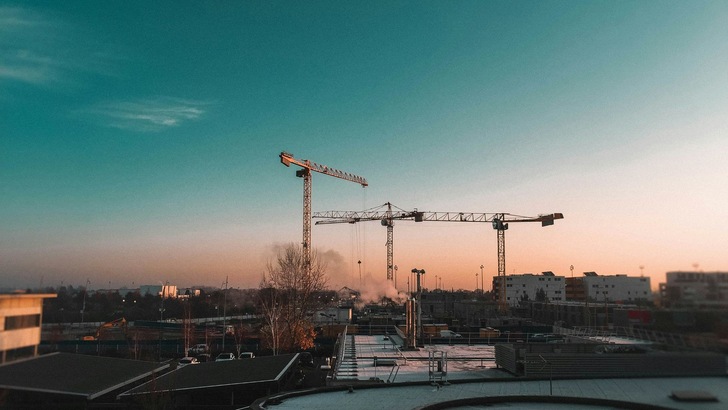 Das Bild zeigt einen großen Baukran vor dem Hintergrund eines farbenfrohen Himmels beim Sonnenuntergang, umgeben von städtischer Bebauung. - © Rubens Nguyen auf Unsplash