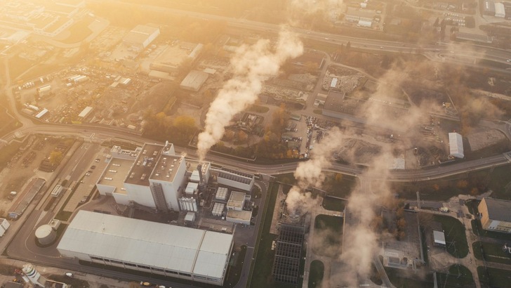 Eine Luftaufnahme zeigt eine Fabrik, aus deren Schornsteinen Rauch aufsteigt. Das umliegende Industriegelände ist ebenfalls zu sehen. - © Marcin Jozwiak, Unsplash