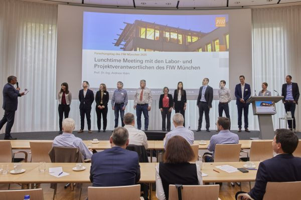 Eine Gruppe von Personen steht während einer Mittagssitzung im FIW München auf der Bühne und blickt auf das Publikum. - © FIW