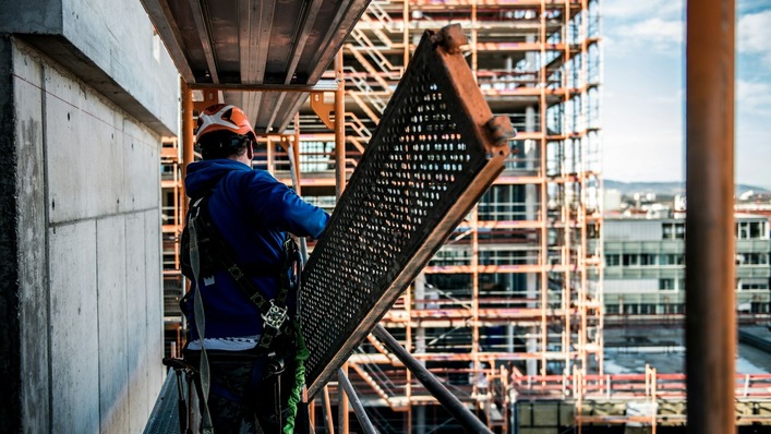 Ein Bauarbeiter in Sicherheitskleidung trägt auf einer Baustelle ein großes Metallgitter. Im Hintergrund ist ein teilweise errichtetes Gerüst sichtbar.