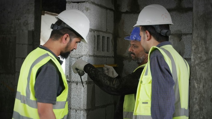 Zwei Bauarbeiter in Schutzausrüstung arbeiten zusammen an einer Mauer auf einer Baustelle.