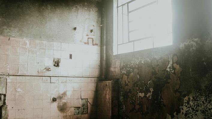 Diese Aufnahme zeigt das Innere eines verlassenen und verfallenen Gebäudes, das durch ein großes Fenster beleuchtet wird. Zu sehen sind beschädigte Fliesen und stark abgenutzte Wände.